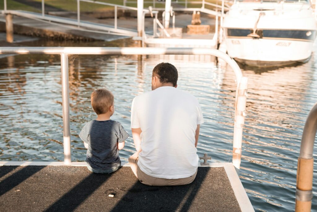 a man and child sitting on a dock looking at a boat